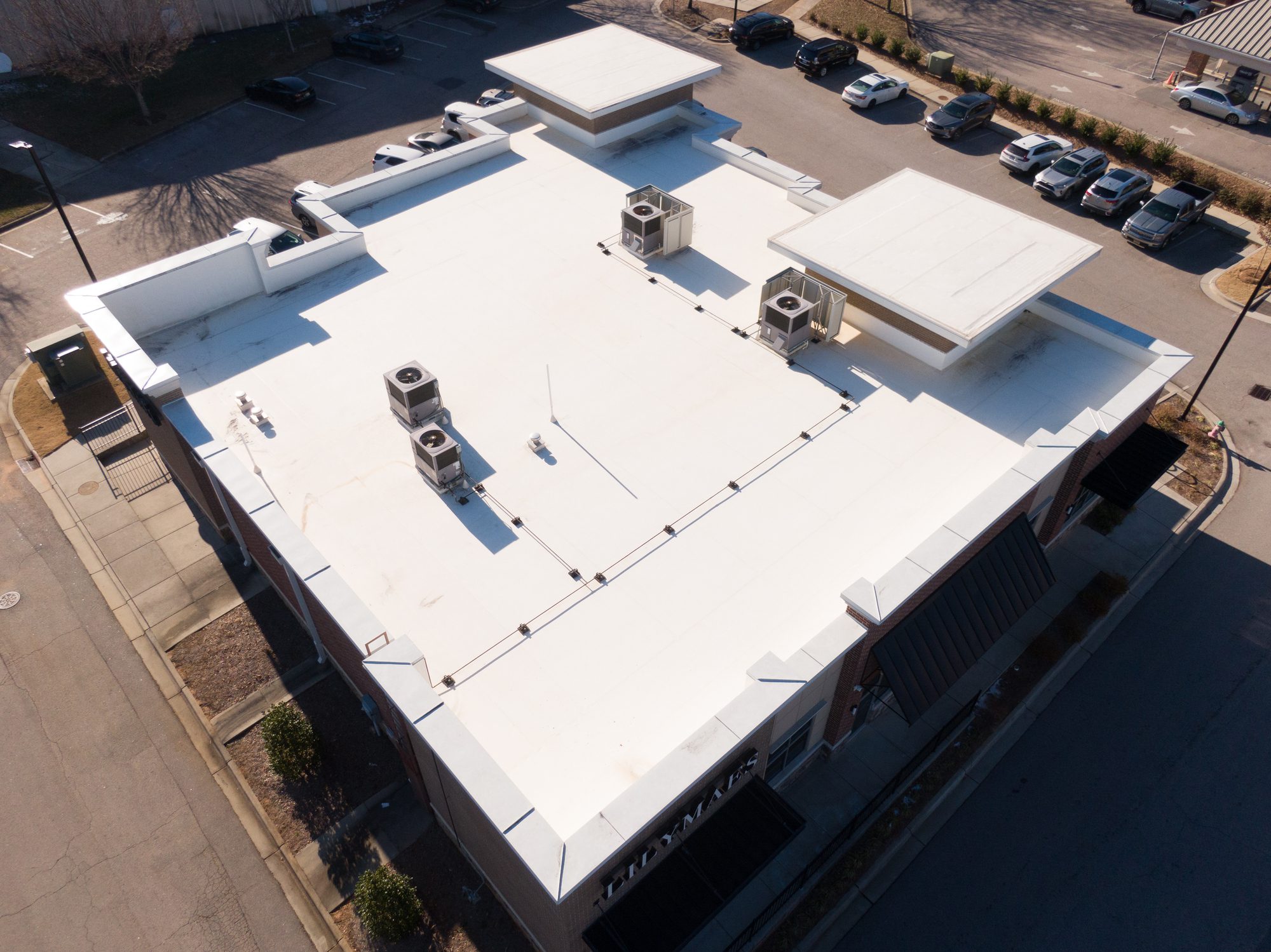 Drone perspective of a flat-roof commercial building in Raleigh, NC. This March 2026 shot showcases an energy-efficient white TPO (Thermoplastic Polyolefin) roofing system, multiple rooftop HVAC units, and integrated drainage and curbing details.