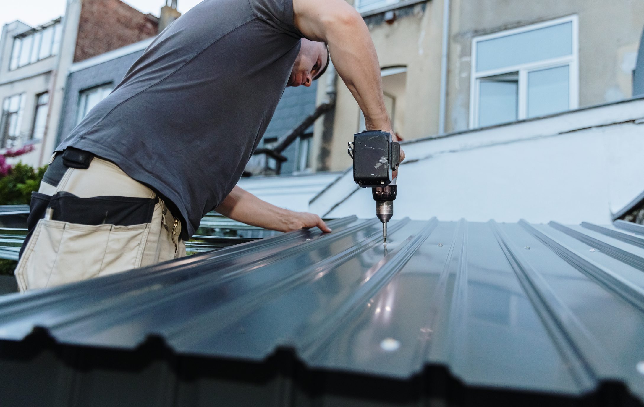A handsome, middle-aged, Caucasian handyman stands sideways on a ladder, installing steel roofing sheets on a metal shed with an electric screwdriver on a summer evening in the backyard. Close-up, bottom-side view. Concept for building a metal shed using technology.