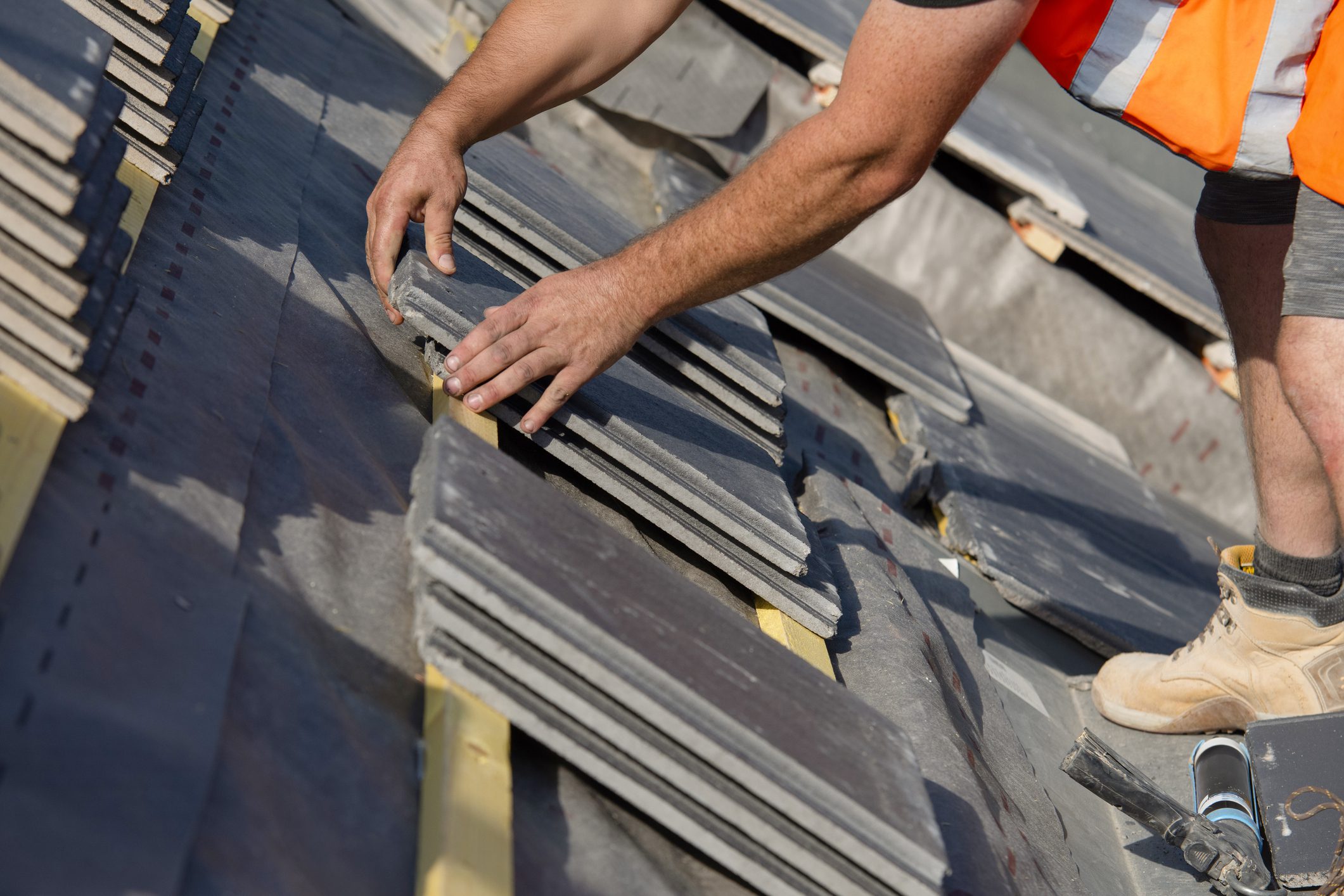 A worker is placing tiles on a roof during daylight. The construction site is busy with materials laid out, and the worker is focused on their task.