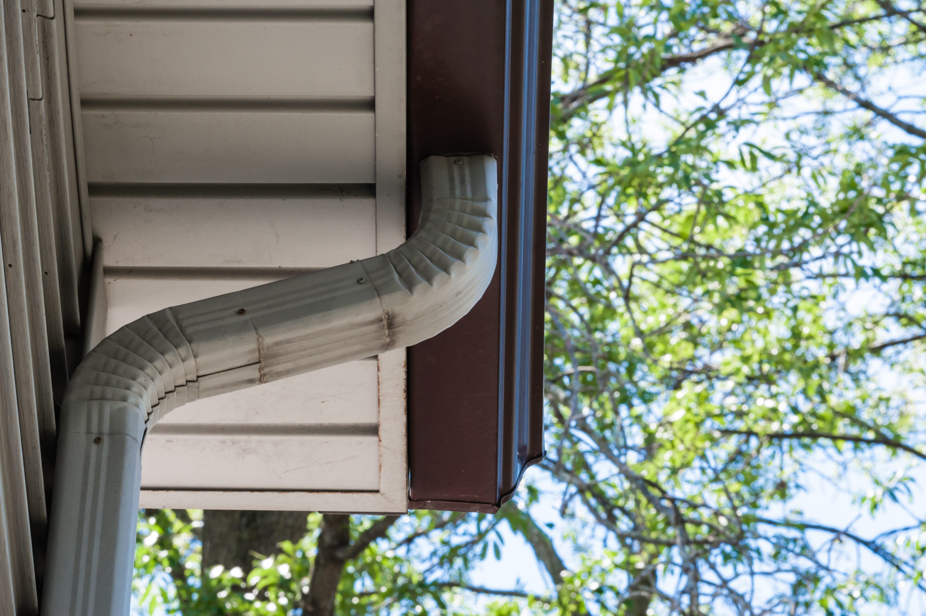 Aluminium gray downspout drain pipe leading down from brown eavestrough, with tree branches and leaves in blurred background.
