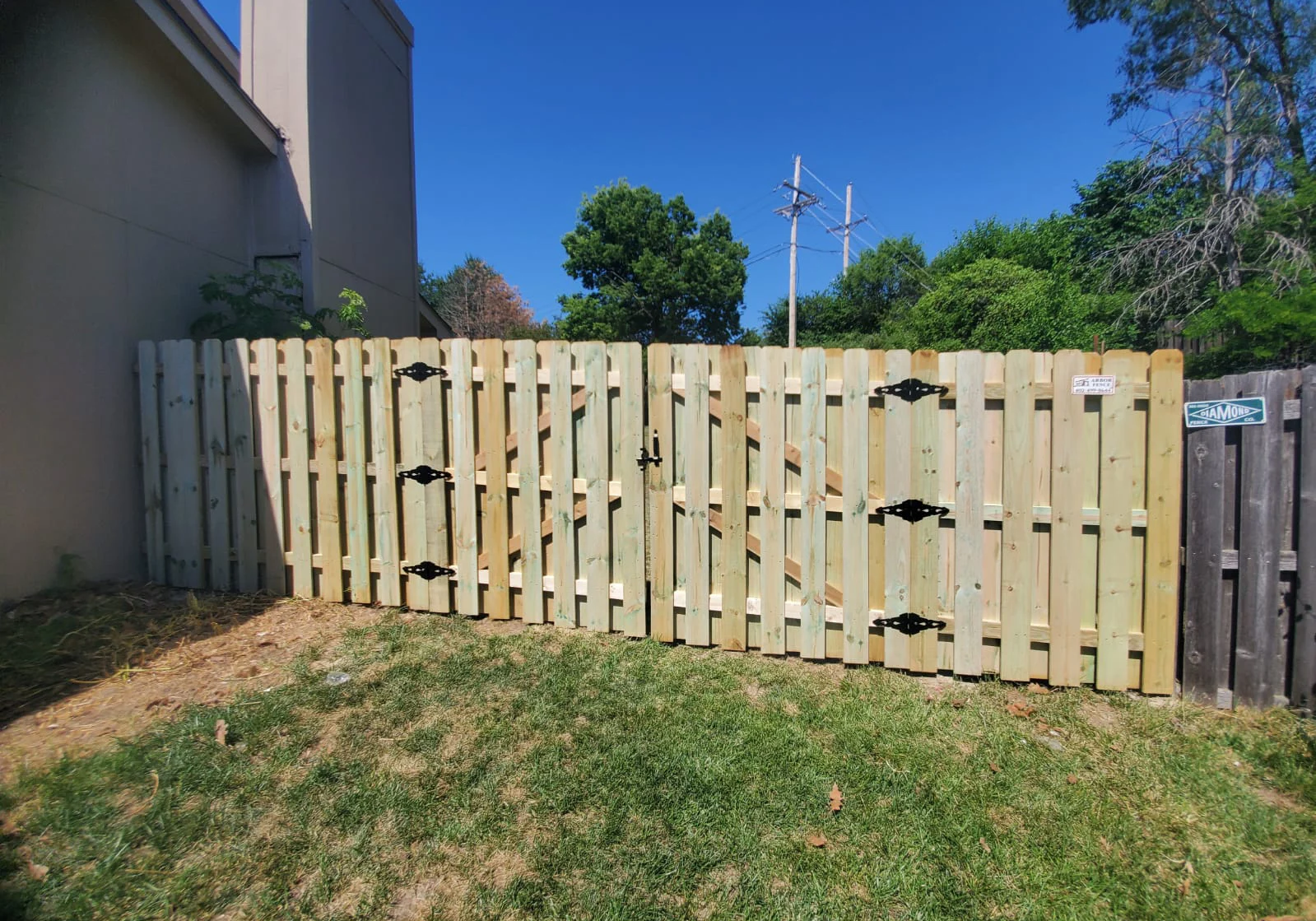 A wooden double gate with black metal hinges and handles is installed between two fences in a grassy yard on a clear, sunny day.