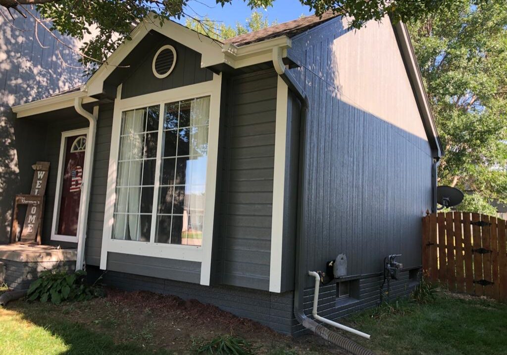 A gray house with white trim, large front window, plants near the foundation, utility meters on the side, and a wooden fence in the background.