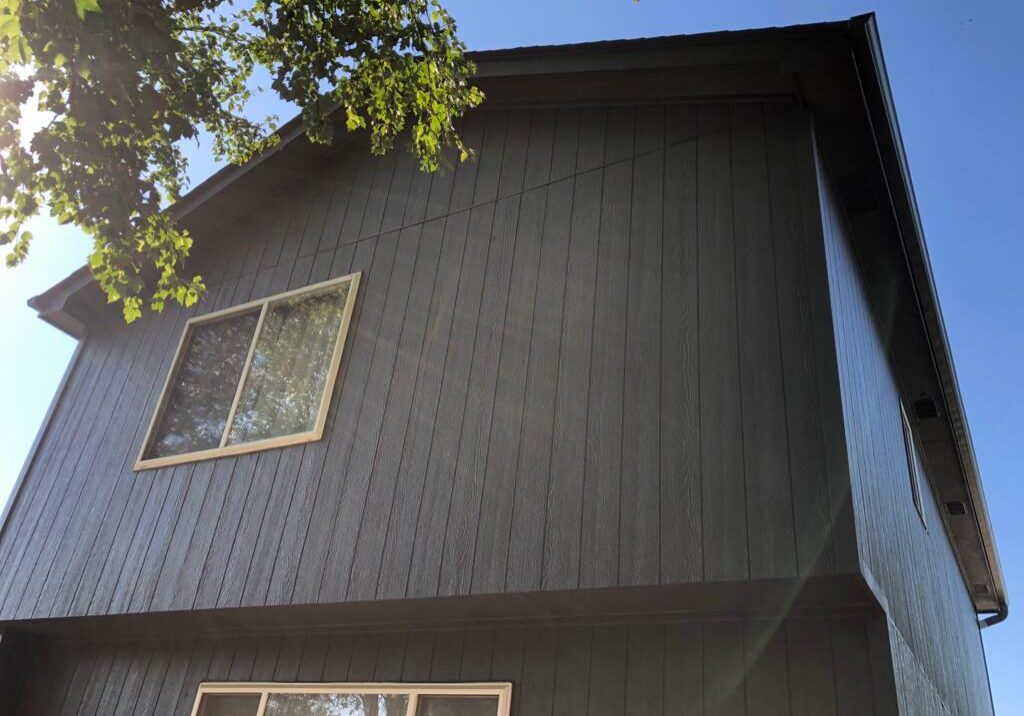 Two-story house with dark wood siding and two rectangular windows, photographed from a low angle on a sunny day with tree branches visible at the top left.